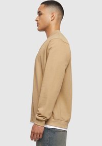 Jeune homme debout de profil, portant une veste beige boutonnée par-dessus une chemise blanche et un jean bleu, sur un fond uni.