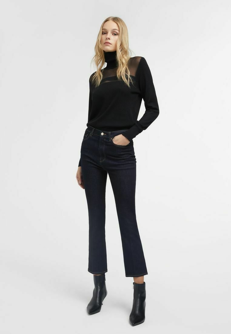 Maglia nera a maniche lunghe con colletto alto e pannello trasparente; abbinata a jeans svasati blu scuro e stivaletti neri alla caviglia. Texture morbide.
