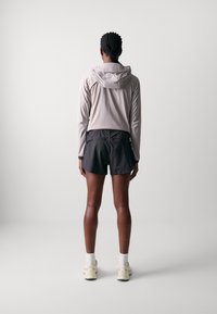 Jasnoszara, długorękawowa hoodie z kapturem, w połączeniu z czarnymi szortami. Model stoi tyłem, nosząc białe skarpetki i żółte trampki.