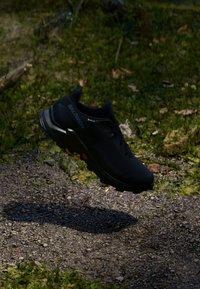 Chaussure de sport noire avec une tige en tissu texturé, semelle robuste et crampons prononcés, affichant un grand logo et un accent rouge.