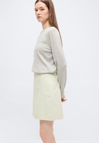 Γυναίκα με μακριά ίσια μαλλιά, που φοράει ανοιχτό γκρι πουλόβερ και κρεμ φούστα σε γραμμή Α, στέκεται μπροστά σε ένα απλό φωτεινό φόντο.