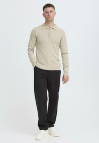 Beige knitted long-sleeve polo sweater with three buttons, ribbed cuffs, and hem, paired with black trousers and white sneakers.