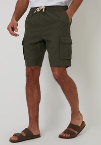 Cargo-Shorts in Olivgrün, mit einem Kordelzugbund, sechs Taschen und einem leichten Stoff. Kombiniert mit braunen Sandalen.