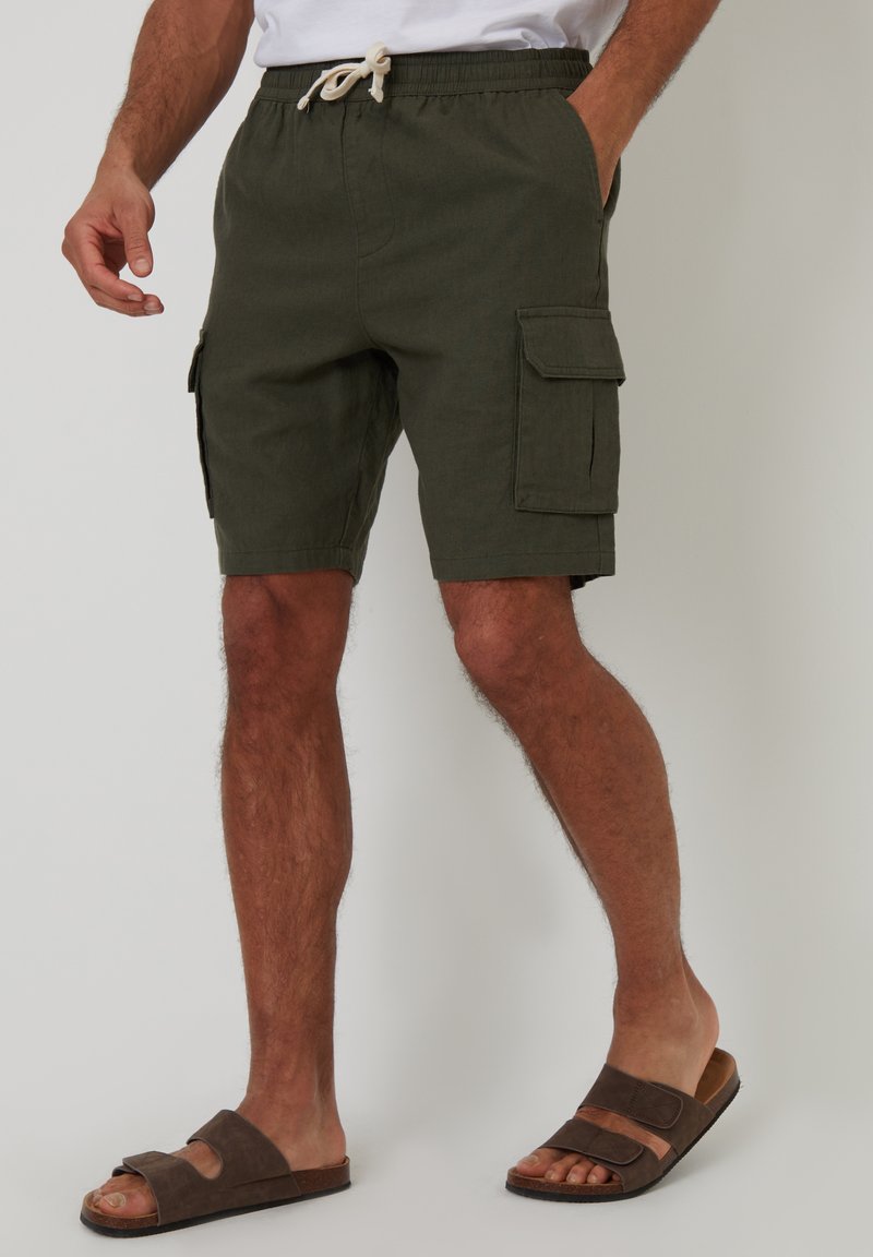 Cargo-Shorts in Olivgrün, mit einem Kordelzugbund, sechs Taschen und einem leichten Stoff. Kombiniert mit braunen Sandalen.
