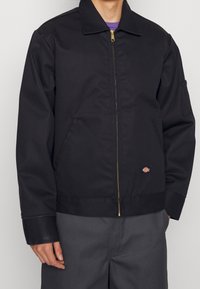 Svart zip-up jacka gjord av slitstarkt tyg. Har en krage, sidofickor och en logotypapplikation på framsidan. Guldzipperdetalj.