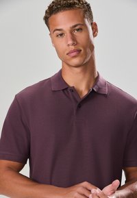 Polo violet foncé avec une surface texturée, manches courtes et une patte à deux boutons. Il présente un col classique et un détail de couture subtil.