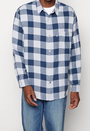 Mężczyzna w niebiesko-białej koszuli w kratę z kieszenią na piersi i jasnoniebieskich dżinsach, stojący na jednolitym tle.