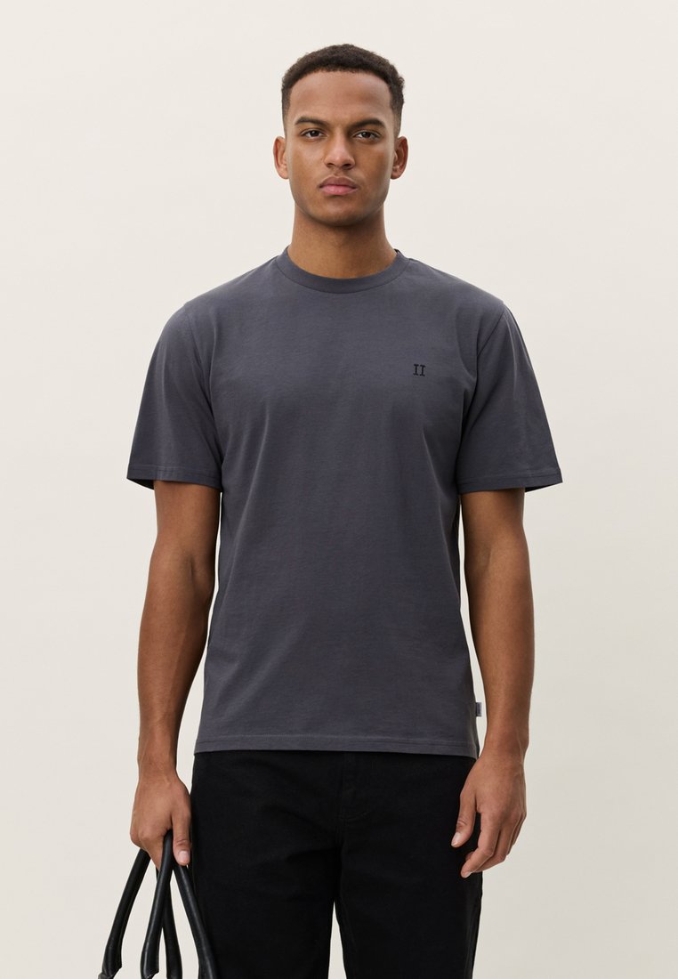 Tumši pelēks kokvilnas T-krekls ar īsām piedurknēm, apaļu izgriezumu, nelielu logotipu krūtīs un brīvu piegriezumu, kombinēts ar melnām biksēm un somu.