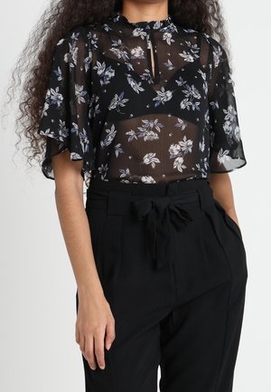 Vrouw draagt een zwarte doorschijnende bloemenblouse met korte, wapperende mouwen en een zwarte broek met hoge taille en een gestrikte riem.