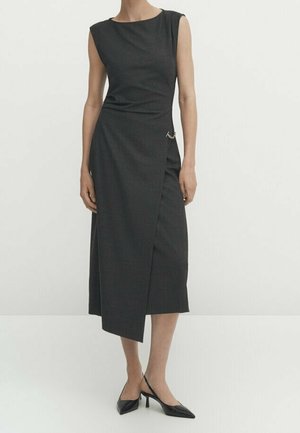 Ærmeløs sort midikjole med asymmetrisk omslagsnederdel og en lille guldfarvet nål, båret med sorte slingback-hæle med spids tå.