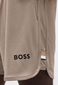 Μπεζ σορτς με κυρτό τελείωμα, πλαϊνή ραφή με μαύρες, λευκές και καφέ ρίγες και το λογότυπο "BOSS" τυπωμένο μαύρο κοντά στο τελείωμα.