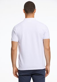 Polo shirt bianco a maniche corte con vestibilità rilassata e aperture laterali, visto da dietro. Tessuto morbido con un design pulito e minimale.