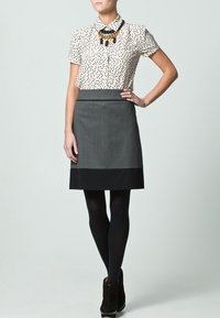 Beige Bluse mit Punkten und kurzen Ärmeln, kombiniert mit einem grauen Bleistiftrock mit schwarzem Saum, ergänzt durch schwarze Strumpfhosen und Stiefeletten.