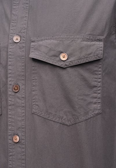 Γκρι ύφασμα πουκάμισου με μπροστινή τσέπη με καπάκι, ασφαλισμένη με ένα μόνο καφέ κουμπί με τέσσερις τρύπες και κάθετη σειρά από αντίστοιχα κουμπιά.