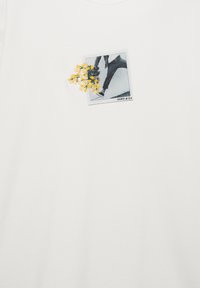 Weißes Baumwoll-T-Shirt mit einem schwarz-weißen Fotodruck und gelber Blumenstickerei, platziert im linken Brustbereich.