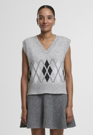 Gilet en laine grise avec un décolleté en V, des manches courtes et un motif en losange noir sur la poitrine. Porté sur une jupe grise évasée.