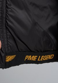 Forro negro de una chaqueta con dobladillo elástico. Presenta un logo amarillo "PME LEGEND" y un emblema de ala. Textura suave, diseño funcional.