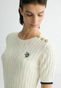 Kreemist soonest kootud sviit lühikeste varrukatega, millel on tumesinised ribasidemed, broderi logo ja kolm dekoratiivset nööpi õlavarre peal.