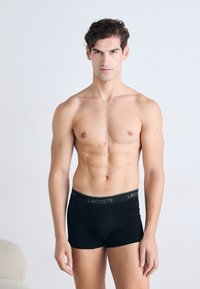 Homem com cabelo curto e encaracolado, sem camisa, vestindo boxers pretos da Lacoste, em pé contra um fundo claro e simples.