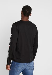Schwarzes Langarm-T-Shirt mit Rundhalsausschnitt und weißem "TOMMY HILFIGER"-Logo entlang des linken Ärmels, kombiniert mit blauen Jeans.