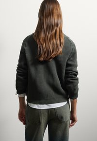 Maglione a coste verde scuro con scollo rotondo e maniche arrotolate, abbinato a jeans di denim azzurri. La texture è visibile nel tessuto a maglia.