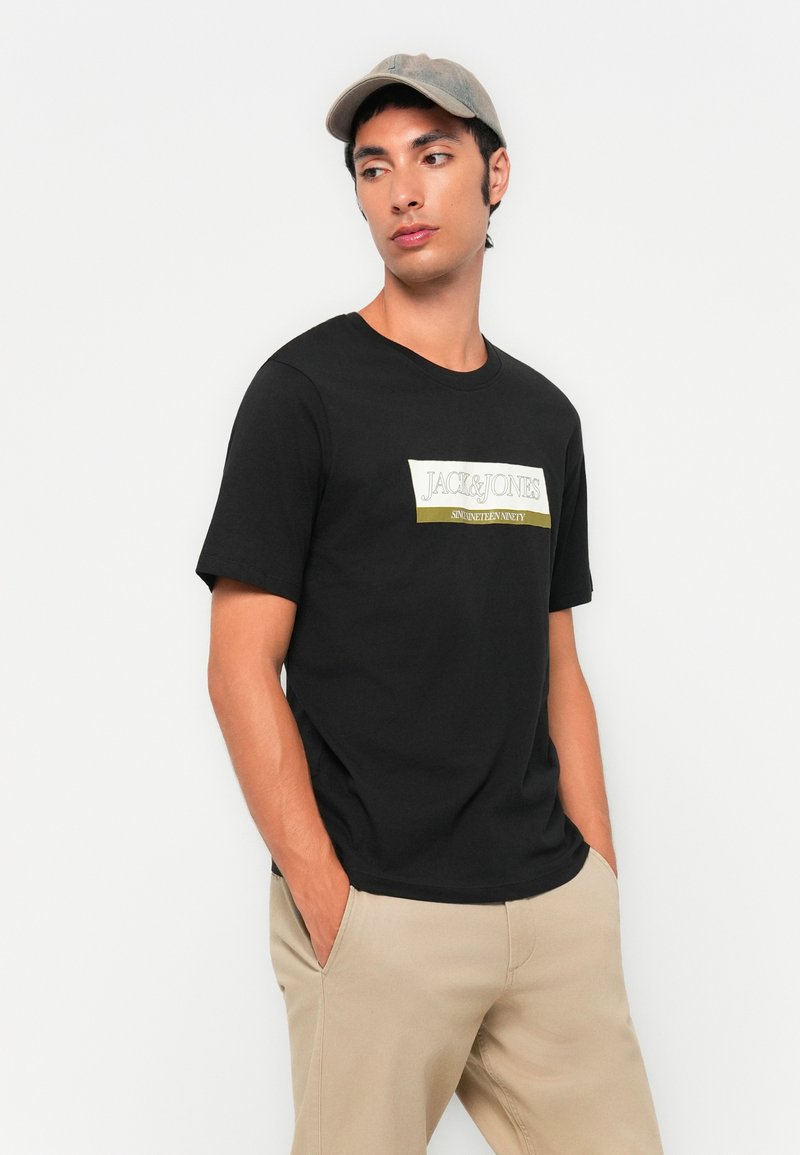 T-shirt en coton noir avec un logo graphique blanc et olive sur le devant. Le modèle porte un pantalon beige et une casquette gris clair.