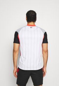 Sportshirt aus weißem Stoff mit vertikalen schwarzen Streifen, grauen und roten Akzenten an den Schultern und tailliertem Design. Kurze Ärmel und Rundhalsausschnitt.