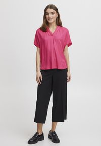 Blusa rosa a maniche corte con scollatura a V e motivo testurizzato, abbinata a pantaloni neri cropped e scarpe nere. Design semplice e moderno.