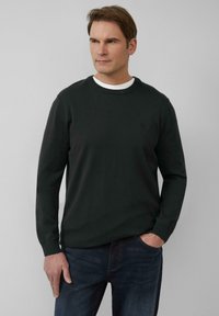 Donkergroene trui met lange mouwen van gebreid materiaal, met een ronde halslijn en strakke manchetten. Draagt over een wit shirt, gecombineerd met een donkere spijkerbroek.