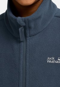 Giacca in fleece blu scuro con colletto alto e zip, caratterizzata dal logo "Jack Wolfskin" in bianco sul petto, superficie texturizzata.
