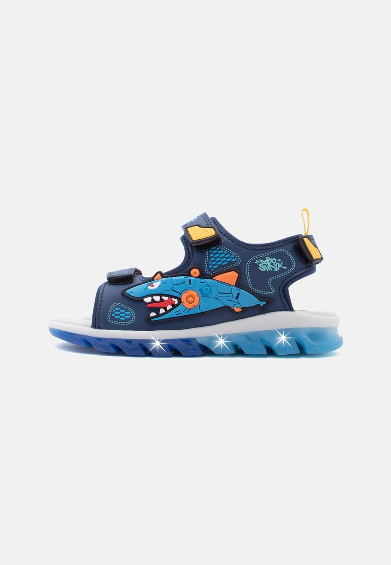 Sandale bleue pour enfants avec motif requin, brides jaunes et semelle bleue lumineuse sur fond blanc.