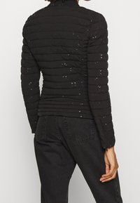 Kobieta w dopasowanej czarnej pikowanej kurtce z subtelnymi błyszczącymi kropkami i czarnych dżinsach, stojąca tyłem z prawą ręką swobodnie opartą przy boku.