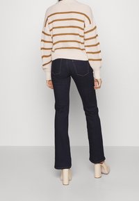 Henkilö, jolla on tummansiniset bootcut-farkut, kerma- ja ruskearaidallinen pusero sekä beiget korkokengät, seisoo selkä kameraan päin yksinkertaisella taustalla.