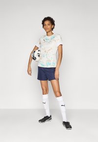 Kortermet fotbaltrøye med mønster i kremfarge med blå detaljer, sammen med marineblå shorts. Sorte fotballsko og hvite sokker med logoer.