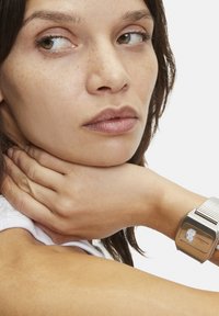 Silverfärgad armbandsklocka med fyrkantig urtavla och mesharmband. Närbild av klockan på ett handled bredvid en hand med neutrala hudtoner.