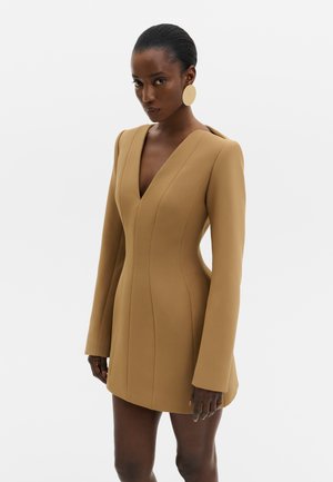 Femme portant une robe blazer cintrée couleur camel avec manches longues et décolleté profond en V, accessoirisée de grandes boucles d’oreilles rondes beiges.
