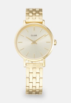 Montre-bracelet en métal doré avec un cadran soleillé doré clair, présentant de fines index horaires et un bracelet en maille poli. Nom de la marque "CLUSE" affiché.
