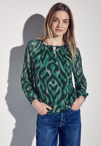Grüne gemusterte Bluse mit einem Schlüssel-Ausschnitt, langen Ärmeln und lockerer Passform, kombiniert mit blauen Jeans. Aus weichem, leichtem Stoff.