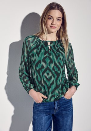 Blusa verde a fantasia con scollatura a chicco, maniche lunghe e vestibilità rilassata, abbinata a jeans blu. Tessuto morbido e leggero.