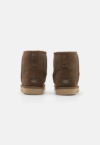 Bottes ankle en daim marron avec une doublure douce, semelles en caoutchouc plates et logo UGG à l'arrière. Présente un design simple et dépouillé.