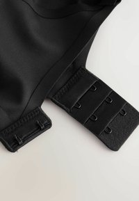 Čierny remienok na podprsenku z látky s tromi radmi háčikov a očiek, rozložený na svetlom povrchu.