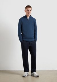 Sweater zippé bleu marine avec une texture douce, associé à un pantalon foncé et des baskets grises. Le pull arbore un petit logo sur la poitrine.