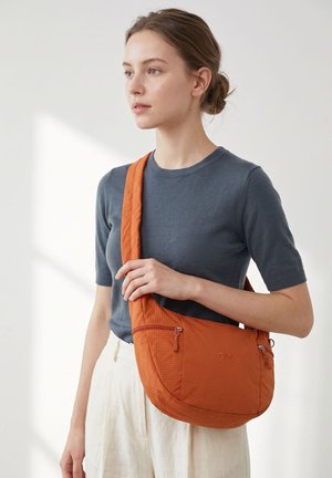 Junge Frau mit Dutt, die ein blaues T-Shirt und weiße Hose trägt und eine strukturierte orangefarbene Umhängetasche mit Reißverschlusstaschen trägt.