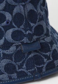 Navy bucket hat made of textured denim features a circular pattern, stitched accents, and a dark blue label with the brand name.