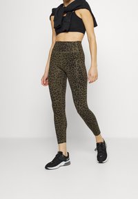 Legginsy z wysokim stanem w oliwkowym kolorze z czarnym wzorem lamparta, wykonane z elastycznego materiału, w połączeniu z czarnym stanikiem sportowym i butami.