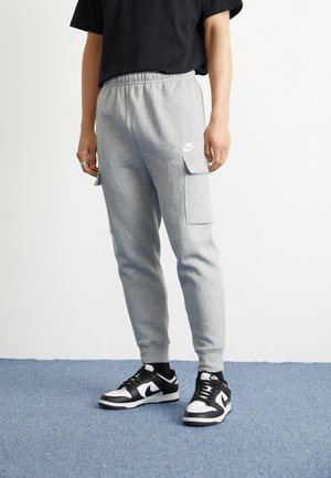 Osoba ubrana w jasnoszare spodnie dresowe Nike z bocznymi kieszeniami typu cargo oraz czarno-białe sneakersy Nike, stojąca na niebieskim dywanie.