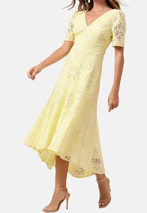 Femme portant une robe midi en dentelle jaune à manches courtes et des sandales à talons hauts couleur nude, debout devant un fond uni.
