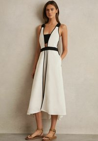 Weißes Midi-Kleid mit schwarzen Akzenten, ausgestattet mit einem tiefen V-Ausschnitt, seitlichen Taschen und einem geflochtenen Taillengurt. Das Model trägt braune, schicke Sandalen.