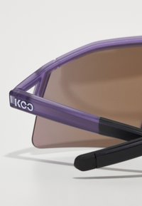 Purple sunglasses with a matte finish, featuring a geometric frame design and gradient brown lenses. Brand logo visible on the arm.