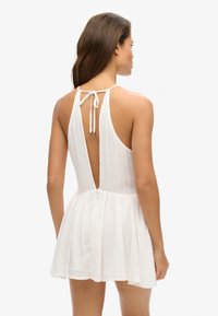 Witte halterjurk met een lage rug, gemaakt van lichtgewicht stof. Beschikt over een uitlopende rok en een striksluiting bij de hals met gouden accenten.